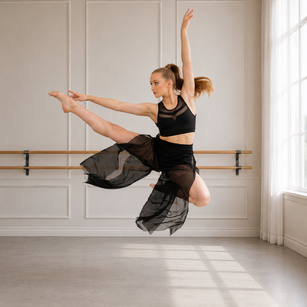 A female dancer in a black dancewear set with sheer mesh panels and a flowing black skirt, captured mid-leap in a bright studio with white paneled walls and a window.