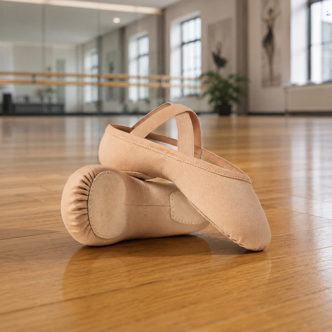 A pair of beige ballet slippers placed on a polished wooden dance studio floor, with a blurred studio interior and windows in the background.