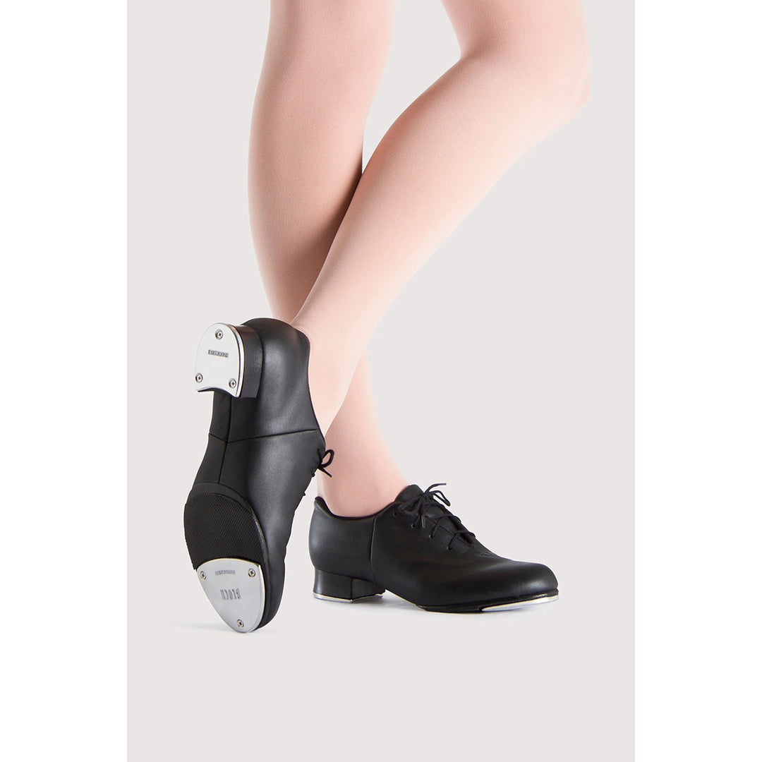 Black leather dance shoes with metal tap heels, shown on a model's feet against a light gray background.