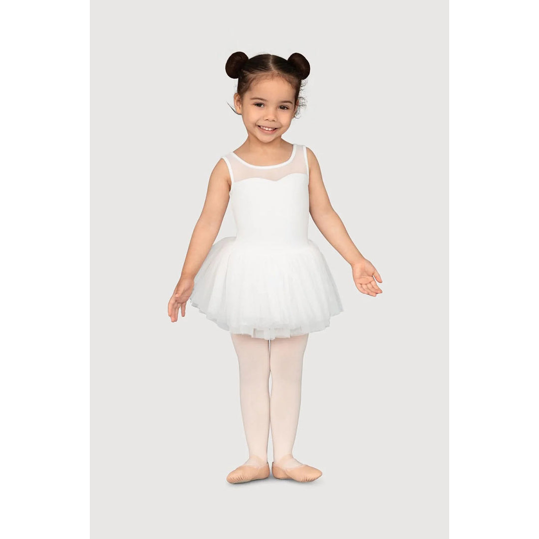 A young girl with her hair in two buns poses in a white sleeveless ballet leotard with a tulle tutu, light pink tights, and ballet slippers against a light gray background.